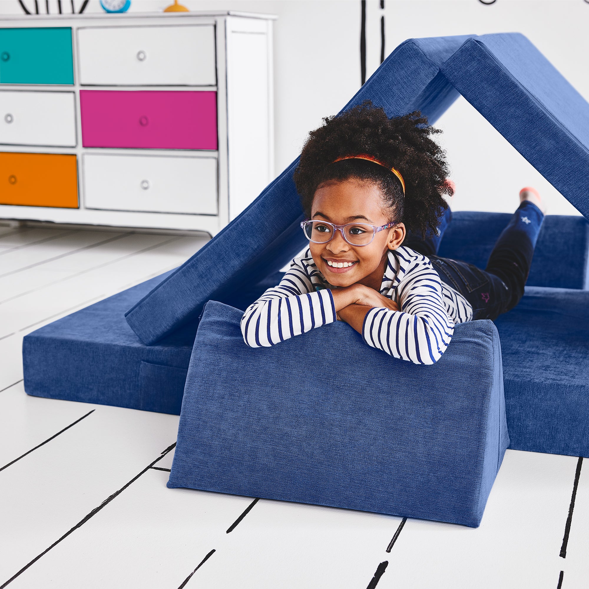 Girl lying on her stomach inside a tunnel configuration of the Yourigami Kids Play Couch in sapphire-caves color with her arms and feet resting on the triangle pieces