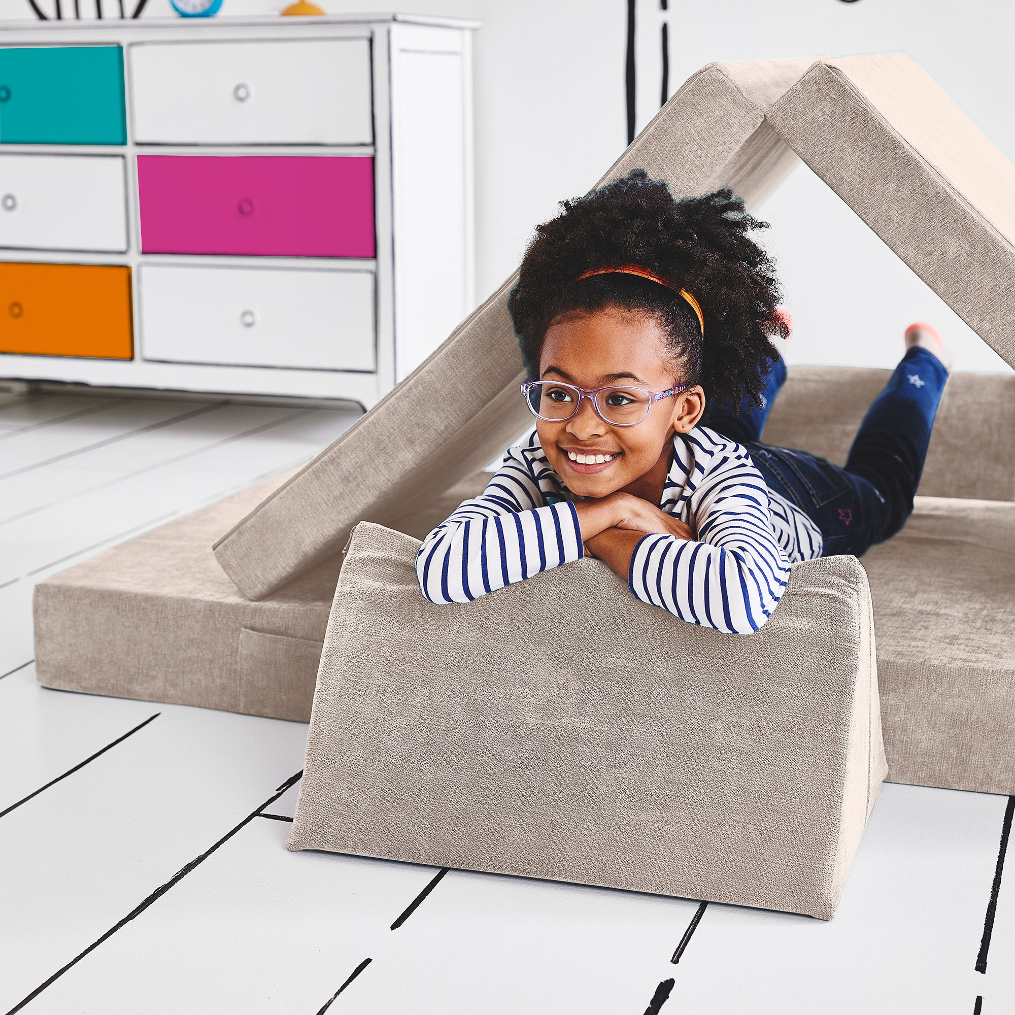 Girl lying on her stomach inside a tunnel configuration of the Yourigami Kids Play Couch in beige-sands color with her arms and feet resting on the triangle pieces