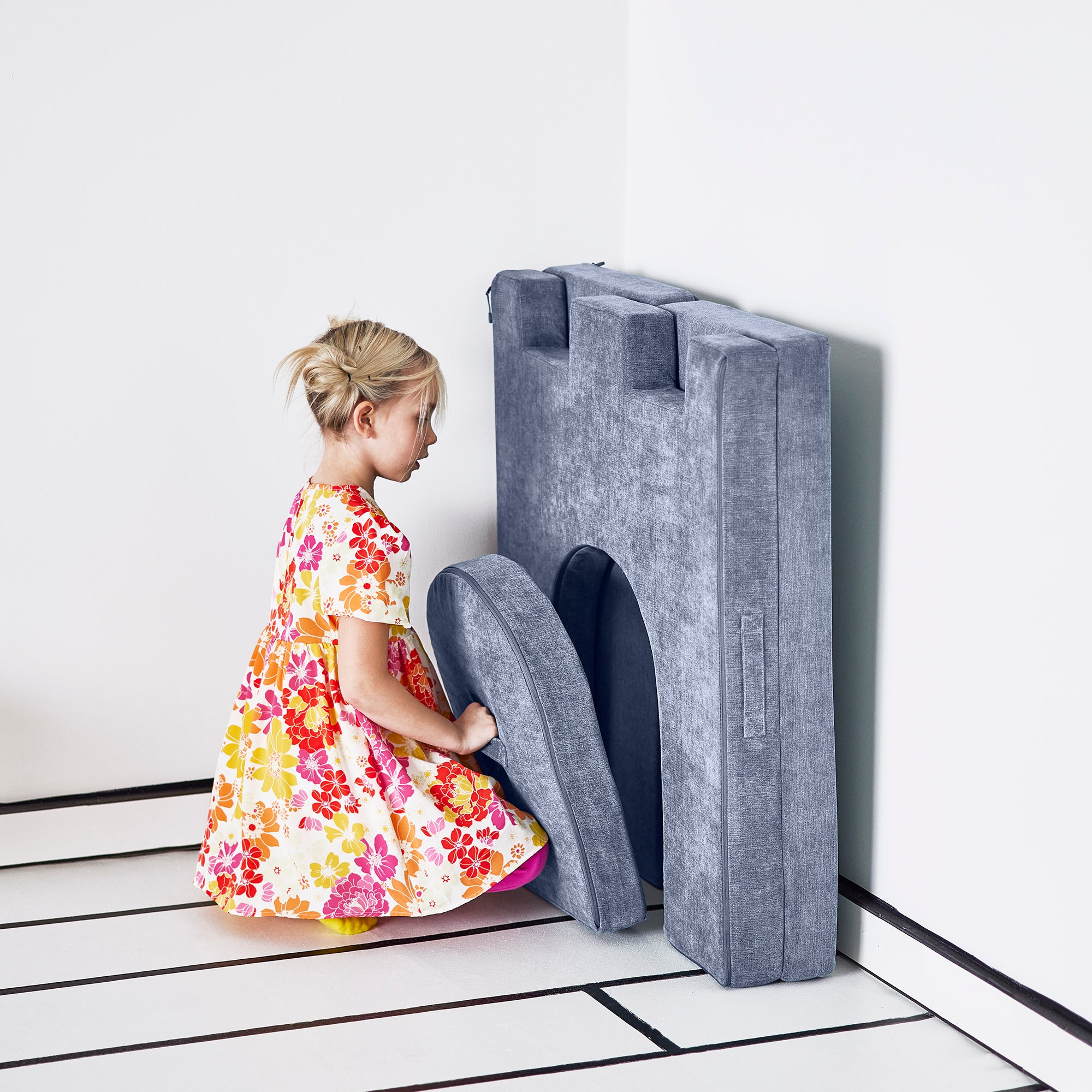 Girl places the drawbridge piece of the Yourigami Kids Play Castle in blue-lagoon color to fit neatly in the folded doorway piece against the wall