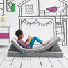 Girl journaling and lying on the Reclined Reader configuration of the Yourigami Kids Play Couch in mountain-gray color with a gaming console in stash pocket