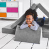 Girl lying on her stomach inside a tunnel configuration of the Yourigami Kids Play Couch in mountain-gray color with her arms and feet resting on the triangle pieces