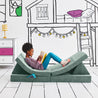 Girl journaling and lying on the Reclined Reader configuration of the Yourigami Kids Play Couch in green-meadows color with a gaming console in stash pocket