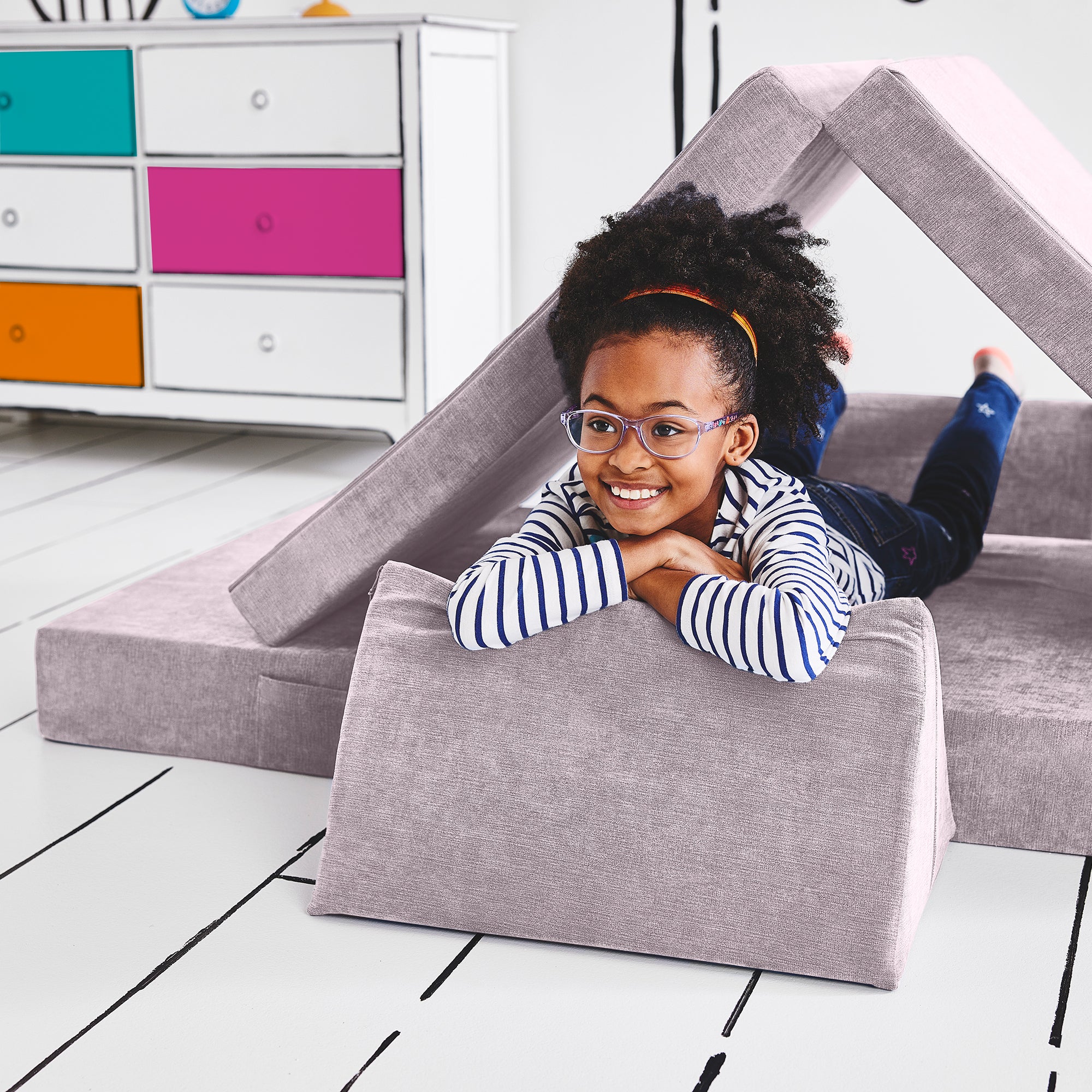 Girl lying on her stomach inside a tunnel configuration of the Yourigami Kids Play Couch in lavender-fields color with her arms and feet resting on the triangle pieces