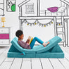 Girl journaling and lying on the Reclined Reader configuration of the Yourigami Kids Play Couch in tropical-teal color with a gaming console in stash pocket