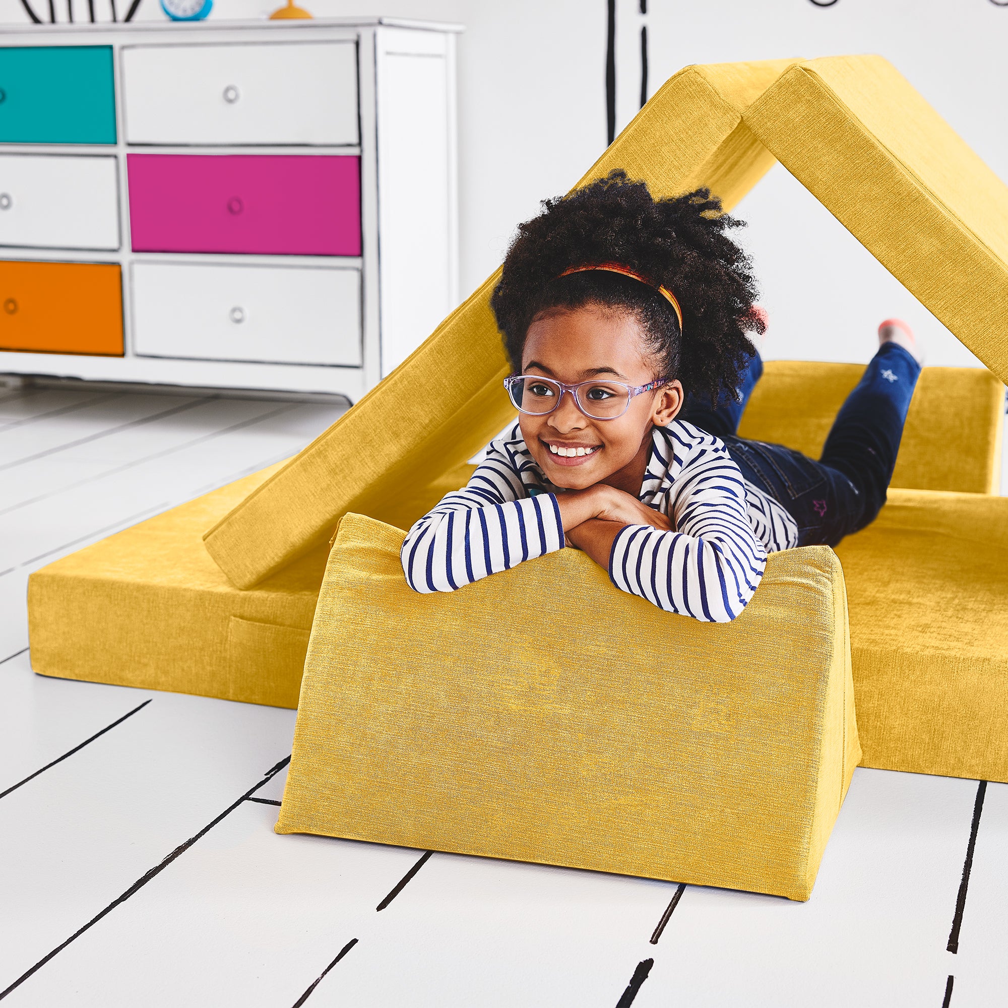 Girl lying on her stomach inside a tunnel configuration of the Yourigami Kids Play Couch in sunflower-yellow color with her arms and feet resting on the triangle pieces